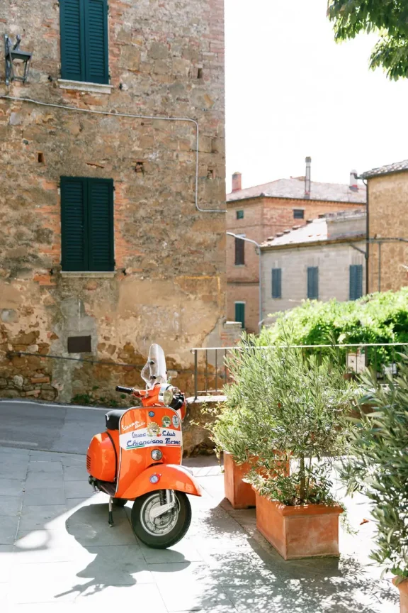 A bright orange scooter parked on a sunlit sidewalk beside large potted plants, with an old stone building and green shutters in the background.